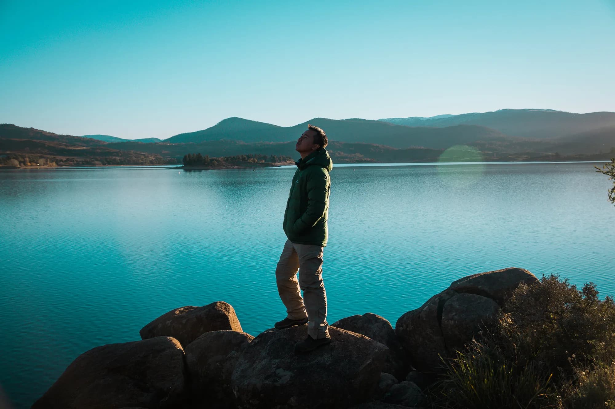 A man standing on rocks by a lake in Australia, looking up at the sky