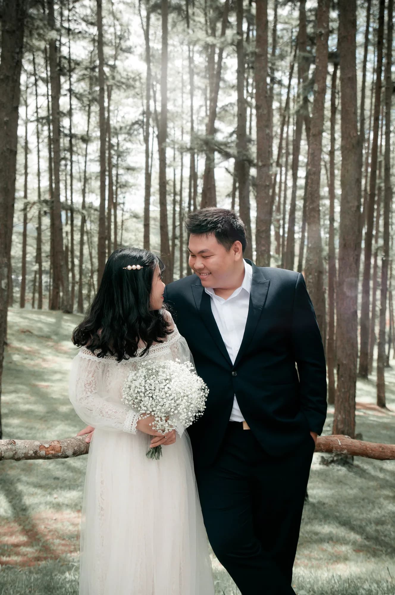 A bride and groom smiling at each other in a forest, holding a bouquet